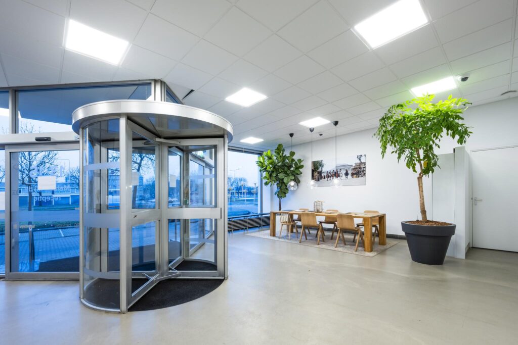 Moderne ontvangsthal aan de Van der Madeweg met draaideur, grote planten en een houten vergadertafel.